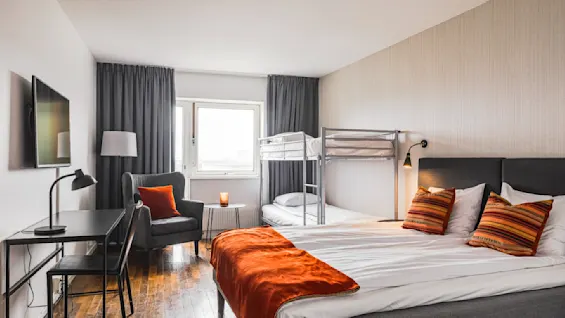 Et koselig hotellrom med en dobbeltseng og køyesenger, med varme oransje aksenter. En stol ved vinduet gir et avslappende hjørne, med et skrivebord og lampe i nærheten for en innbydende atmosfære.