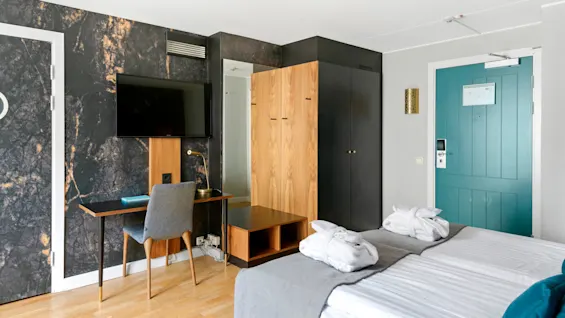 A modern hotel room with twin beds, a desk, TV, and wardrobe, featuring a black marble wall and aqua door.