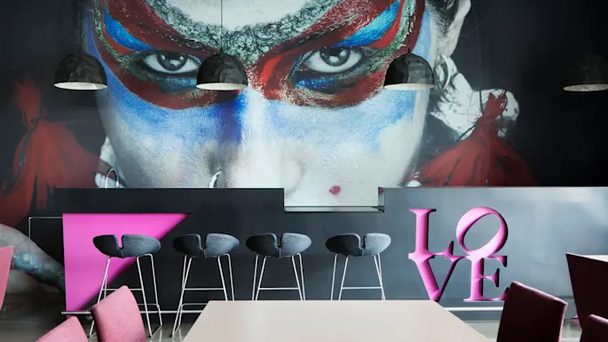 A wall painting of a face with colorful paint surrounds a bar with black stools. Pink letters spell "LOVE". Hanging lights illuminate the scene.