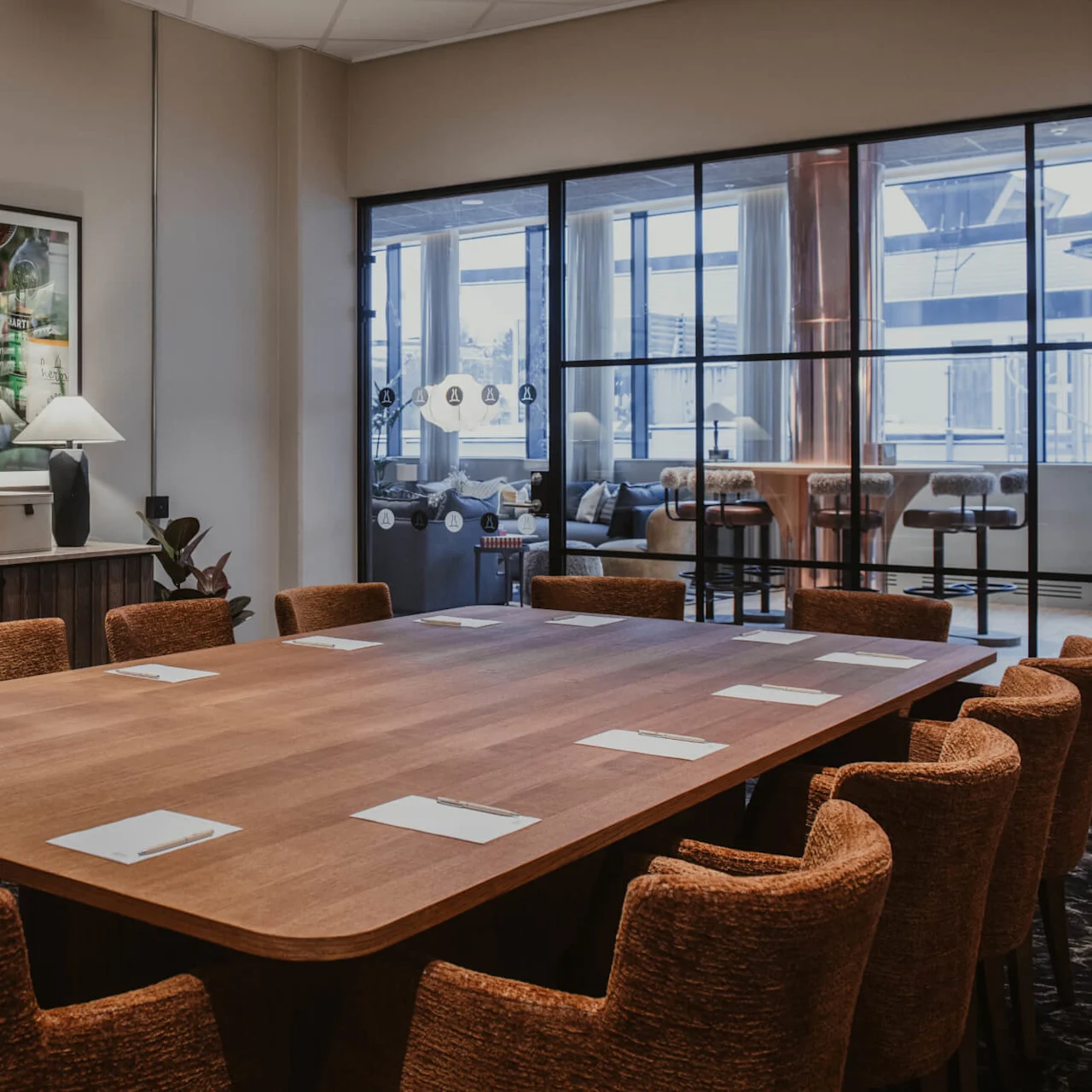 En lang träbord med bruna stolar har anteckningsblock och pennor, placerade i ett modernt konferensrum med inramad konst på väggarna och en glaspanel som visar ett loungeområde.
