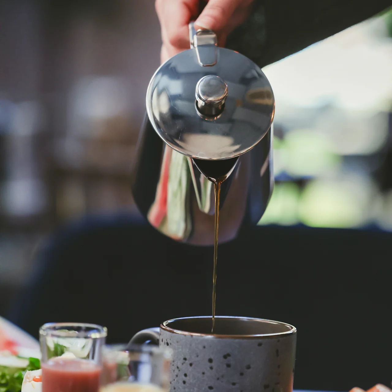 A hand pours coffee from a metal pot into a speckled mug on a table with breakfast items, including juices and vegetables, in a softly blurred background.
