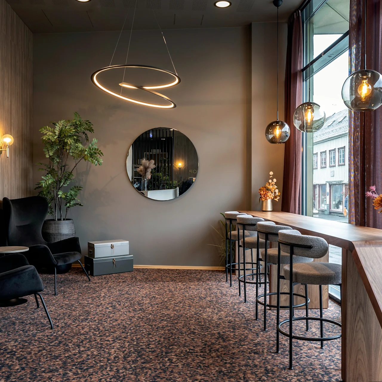 A stylish lounge with dark chairs, circular ceiling lights, a round wall mirror, and a decorative plant. Bar stools line a wooden counter by large windows with street view.