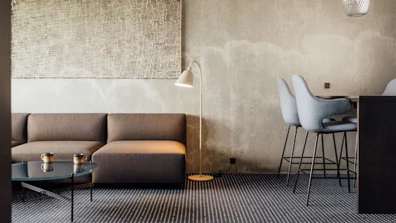 En brun sofa står i en moderne stue med et mønstret vægophæng ovenover. En gulvlampe oplyser, ledsaget af to grå barstole ved en lille bar.