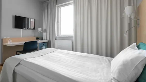 En prydlig hotellrum med en snyggt bäddad säng. En väggmonterad TV och ett skrivbord med en stol är placerade nära ett fönster täckt av ljusa gardiner.