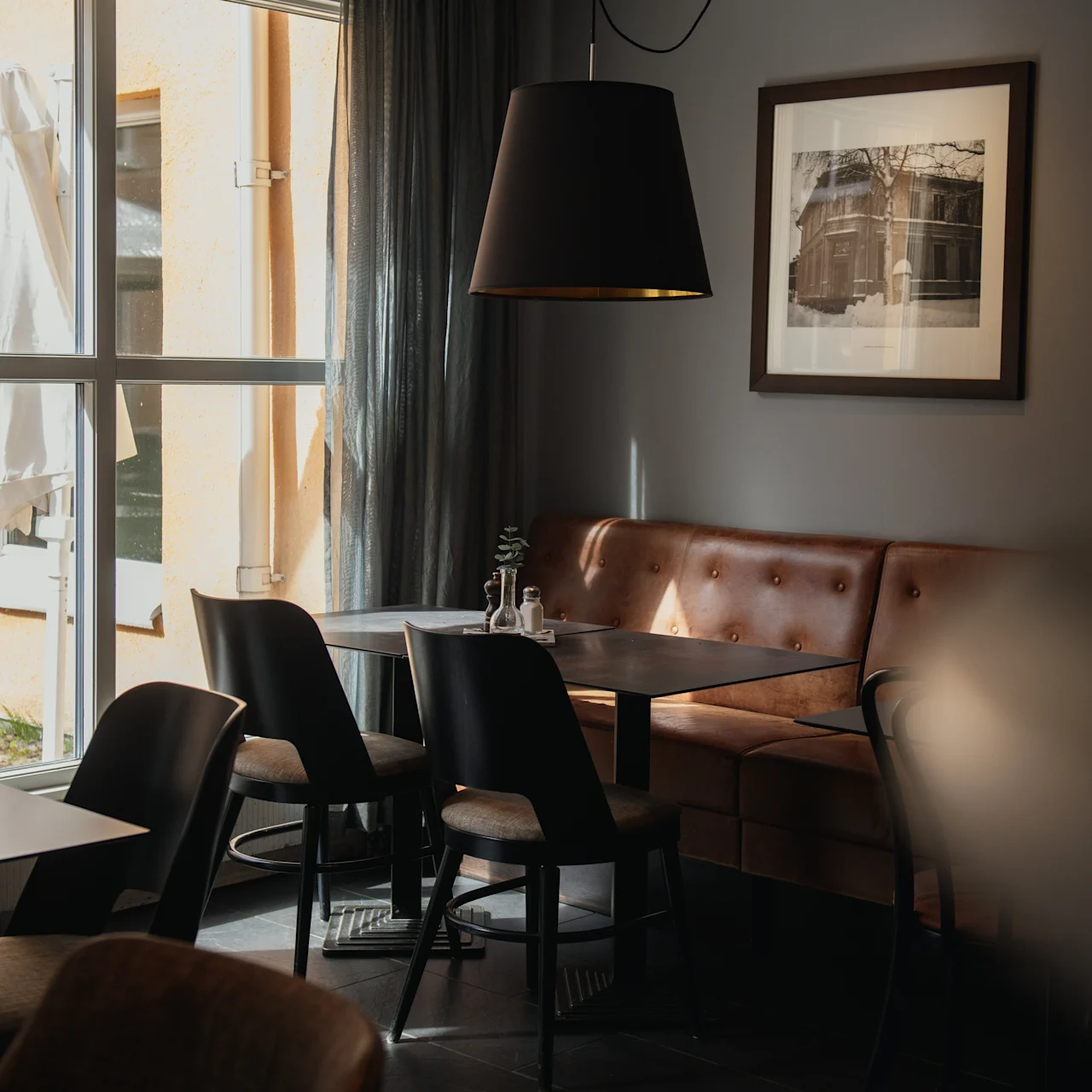 Cozy dining area with a brown leather booth and wooden chairs surrounding a dark table, lit by a ceiling lamp. Two framed pictures hang on dark walls, near large window.