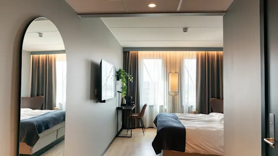 Et moderne hotellrom med en seng, et skrivebord med TV, et stort speil og et vindu med gardiner.