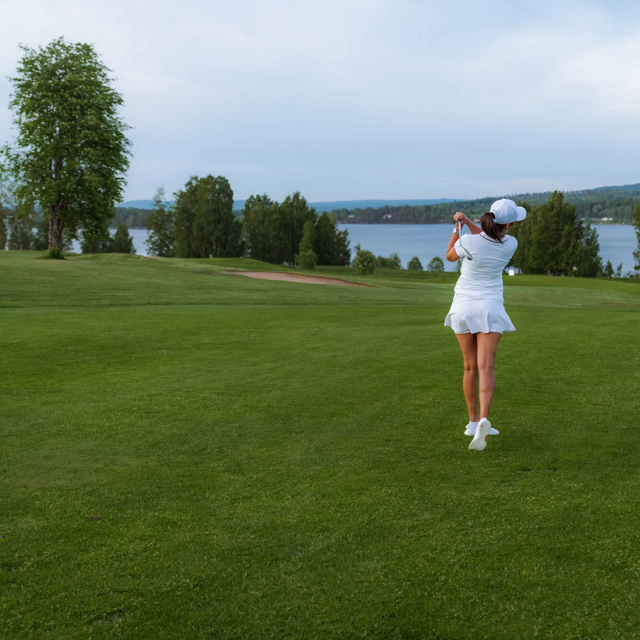 En golfspiller i hvitt antrekk svinger en kølle på en gressbane nær en innsjø, omgitt av trær under en skyet himmel.