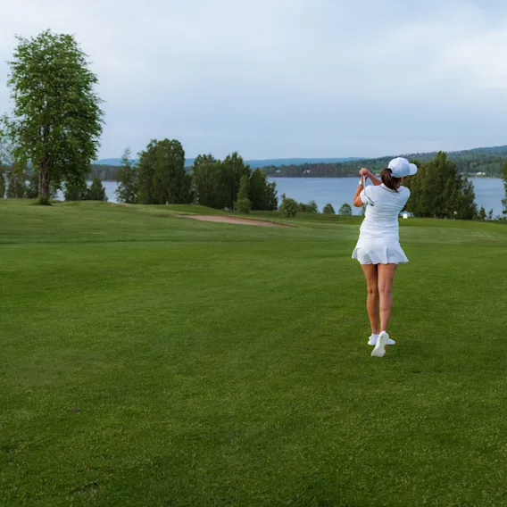 En golfspiller i hvitt antrekk svinger en kølle på en gressbane nær en innsjø, omgitt av trær under en skyet himmel.