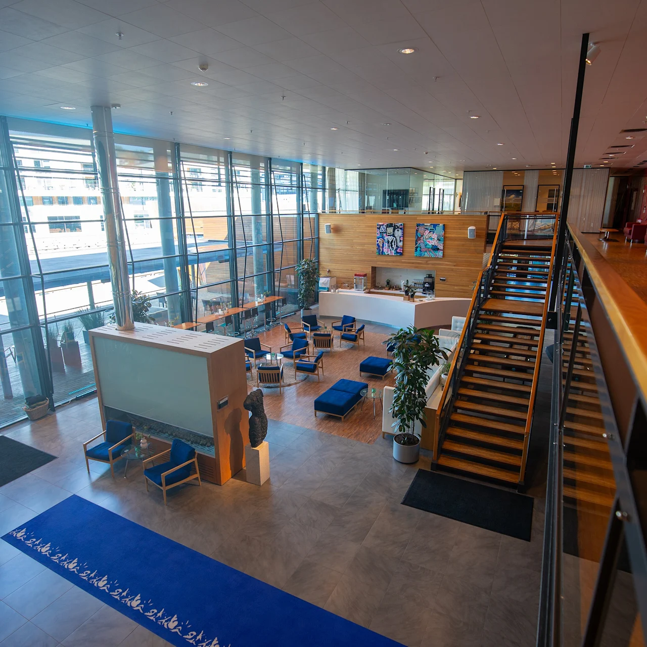Stor åpen lobby med tretrapp som går opp til rødteppebelagt overetasje. Blå sitteplasser og abstrakt kunst på veggene. Glassvinduer viser gate utenfor.