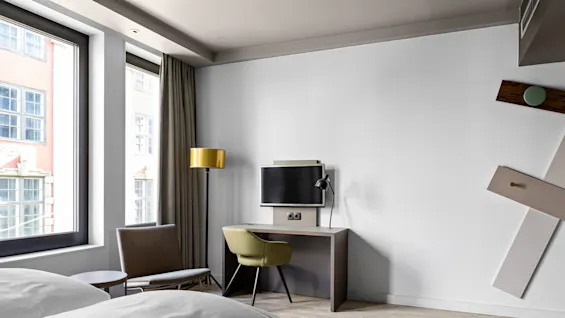A modern hotel room features a bed, desk with a television, and a green chair. A floor lamp stands by the window with curtains, and a decorative wall design.