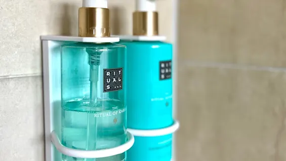Two teal pump bottles labeled "RITUALS," attached to a wall mount in a bathroom setting. Text on the front bottle: "THE RITUAL OF KARMA" and "shampoo & shower gel holy lotus & organic white tea."