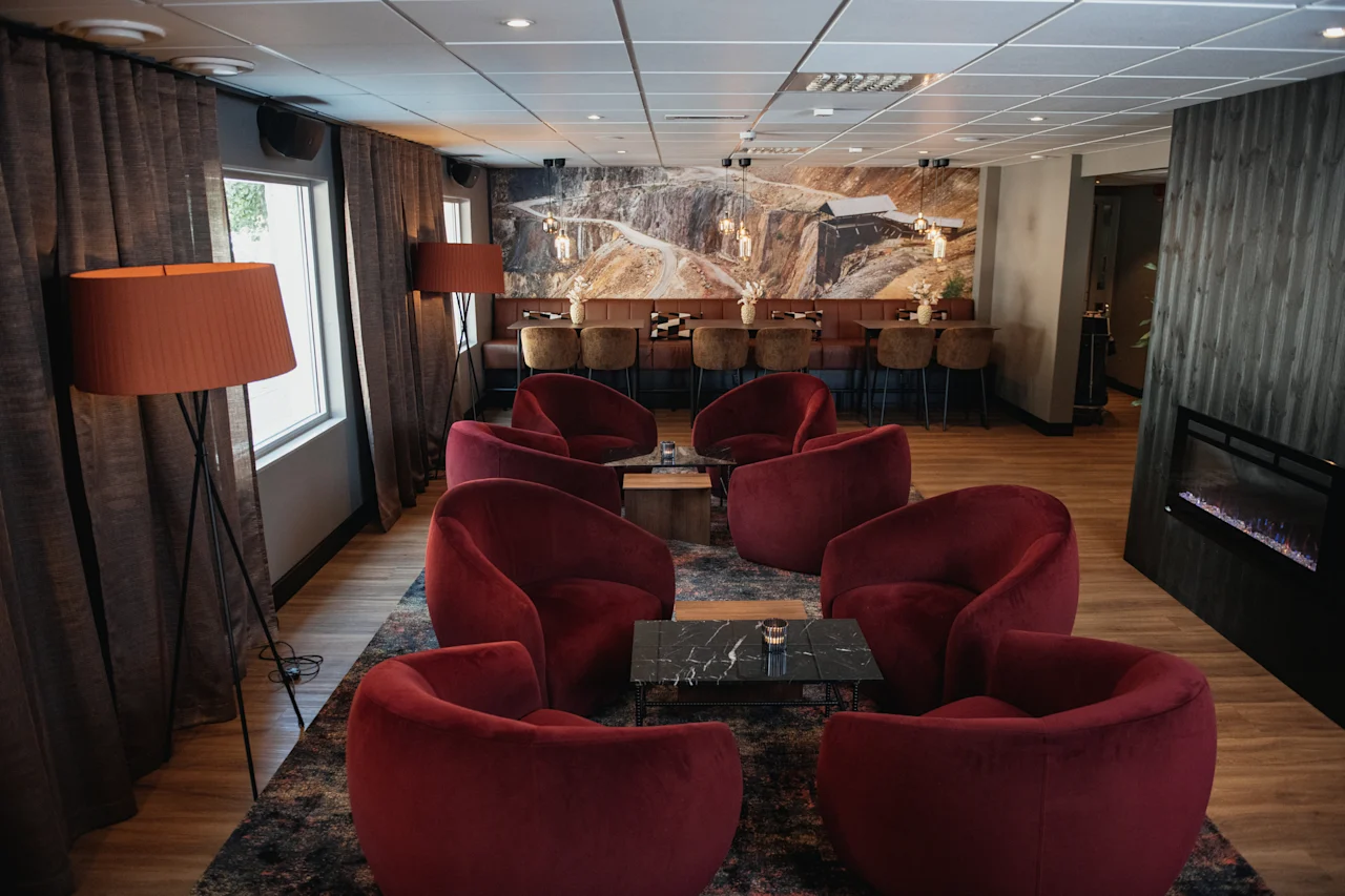 En innbydende hotellounge med komfortable røde lenestoler, små bord og et spiseområde med et stort landskapsmaleri.