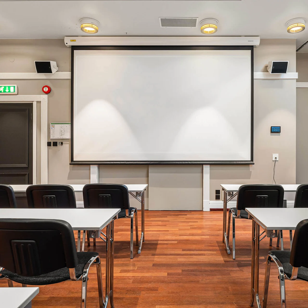 Et lærred til projektion er monteret på en væg i et konferencelokale. Flere stole og borde er vendt mod det. Rummet har trægulv og lysegrå vægge.
