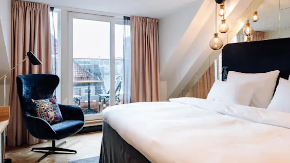 A stylish hotel room features a large bed, a modern armchair with a colorful pillow, and a floor lamp, set against a window overlooking a rooftop terrace.