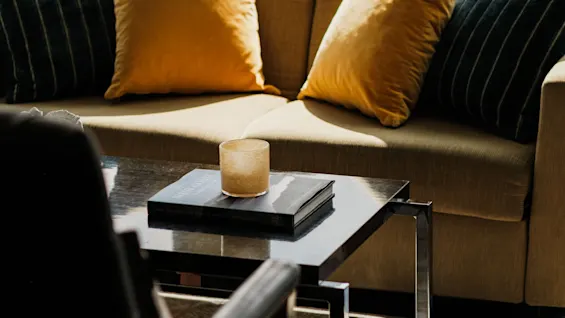 Et hyggeligt hotelloungeområde med en sofa, puder og et sofabord med et lys og en bog, med udsigt over vandet.