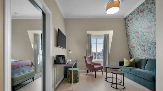 Ett modernt hotellrum med en grön sammetssoffa, blommig tapet och utsikt över en stad genom fönstret.