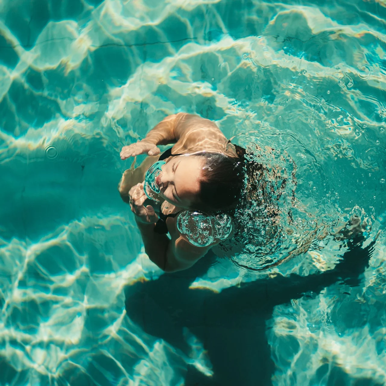 En person svømmer glad i en klarblå pool, med vandsprøjt og bobler omkring ansigtet, og nyder et forfriskende øjeblik.