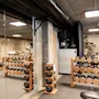 Håndvægte og kettlebells står på trærack i et moderne, rummeligt motionscenter med betonvægge og -loft. Motionscykler og reb er synlige i baggrunden.