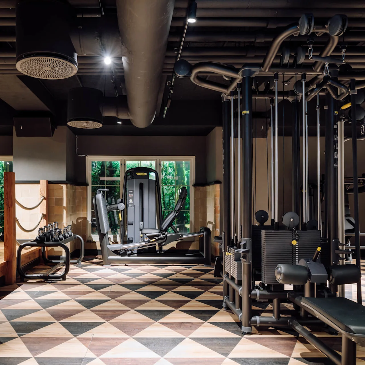 Gym equipment stands idle on patterned floor; kettlebells atop wooden platform; large windows reveal greenery. Black ceiling features air ducts.