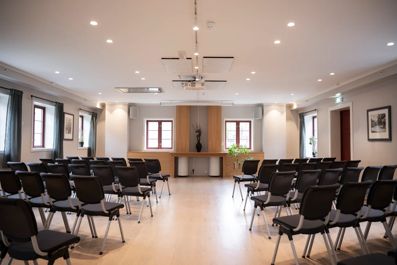 Rows of empty black chairs face a wooden podium with a vase, in a bright room with large windows and framed pictures on light-colored walls.