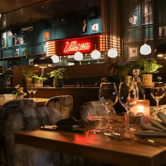 Mysig restauranginteriör med dukade träbord, vinglas, levande ljus och en lysande röd neonskylt.