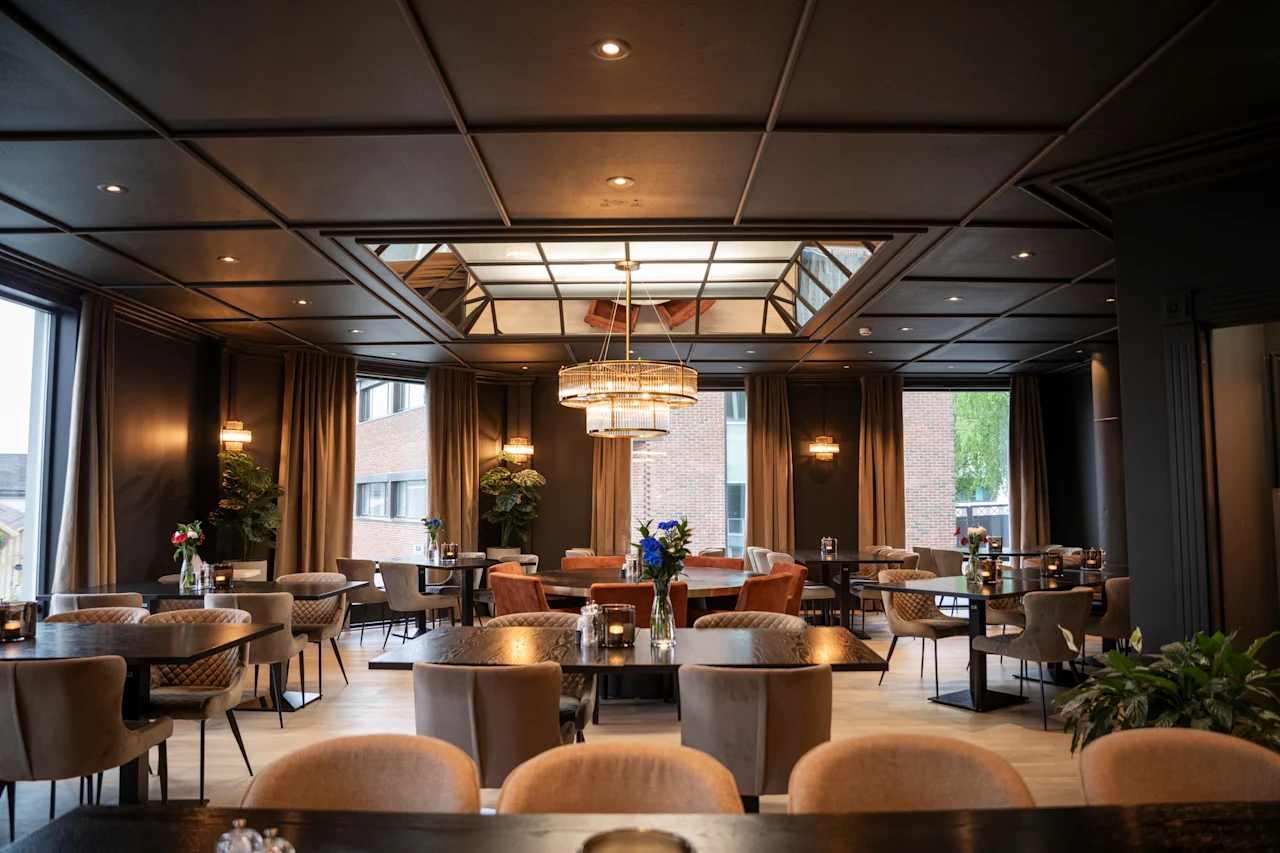 En elegant restaurantspisesal med flere borde dækket til gæster, med store vinduer og en stilfuld lysekrone.