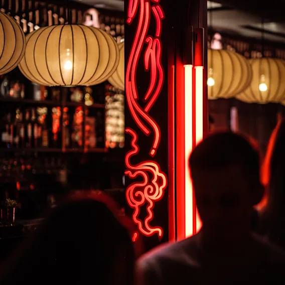 Neonlys kaster et rødt skjær over en svakt opplyst bar med papirlanterner, som skaper silhuetter av hoder i forgrunnen.