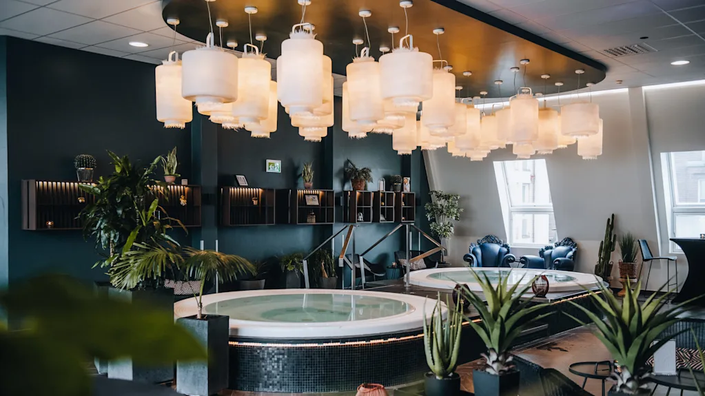 Et rundt boblebad hviler under et tak prydet med lanterner, omgitt av grønne planter og moderne dekor. Miljøet ser ut til å være et luksuriøst, godt opplyst spa-rom.