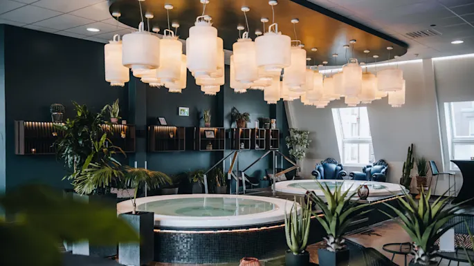 Et rundt boblebad hviler under et tak prydet med lanterner, omgitt av grønne planter og moderne dekor. Miljøet ser ut til å være et luksuriøst, godt opplyst spa-rom.