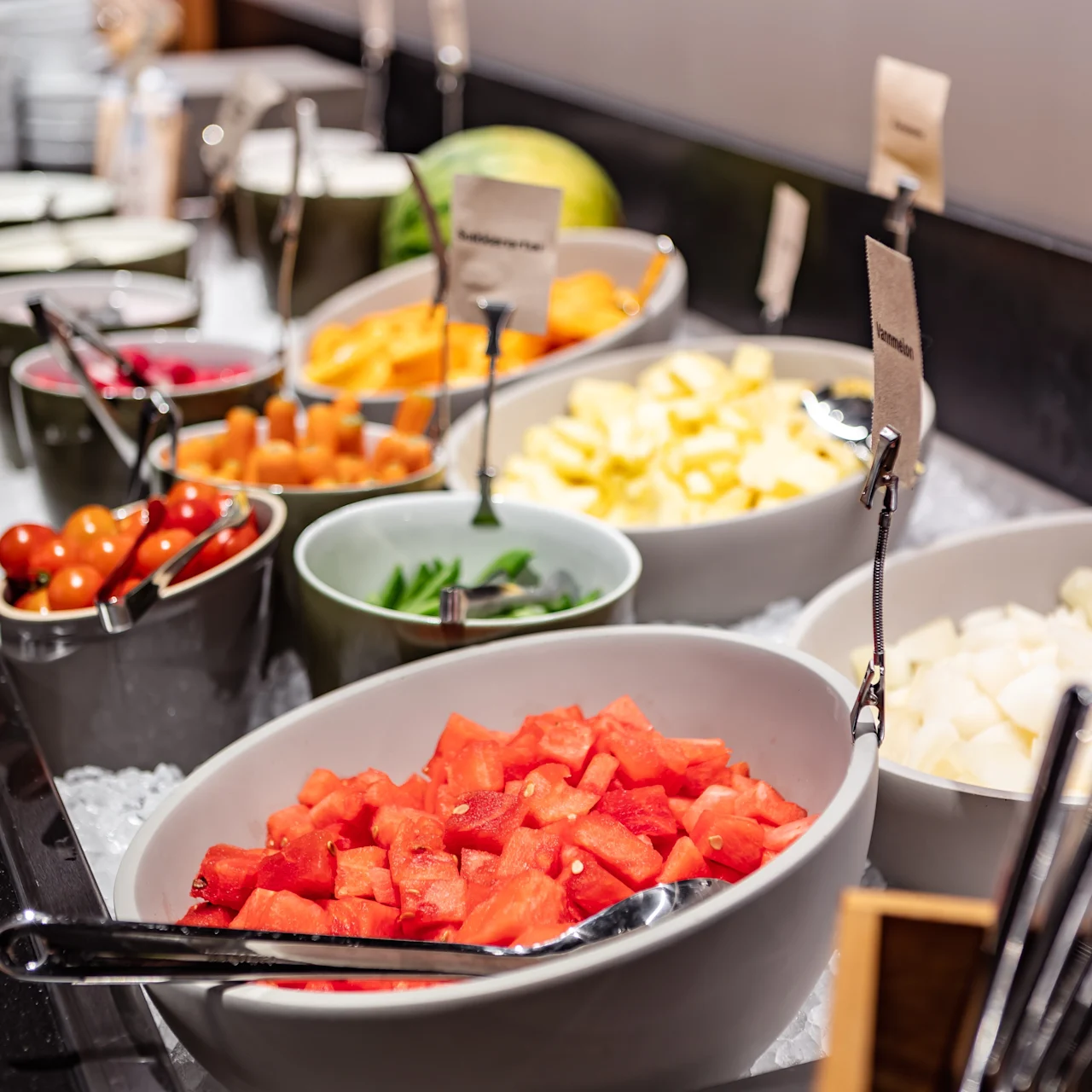 Bowls holding cut fruit and vegetables are displayed on a buffet table. Ice surrounds the containers for freshness. Nearby utensils and small signs label each item.