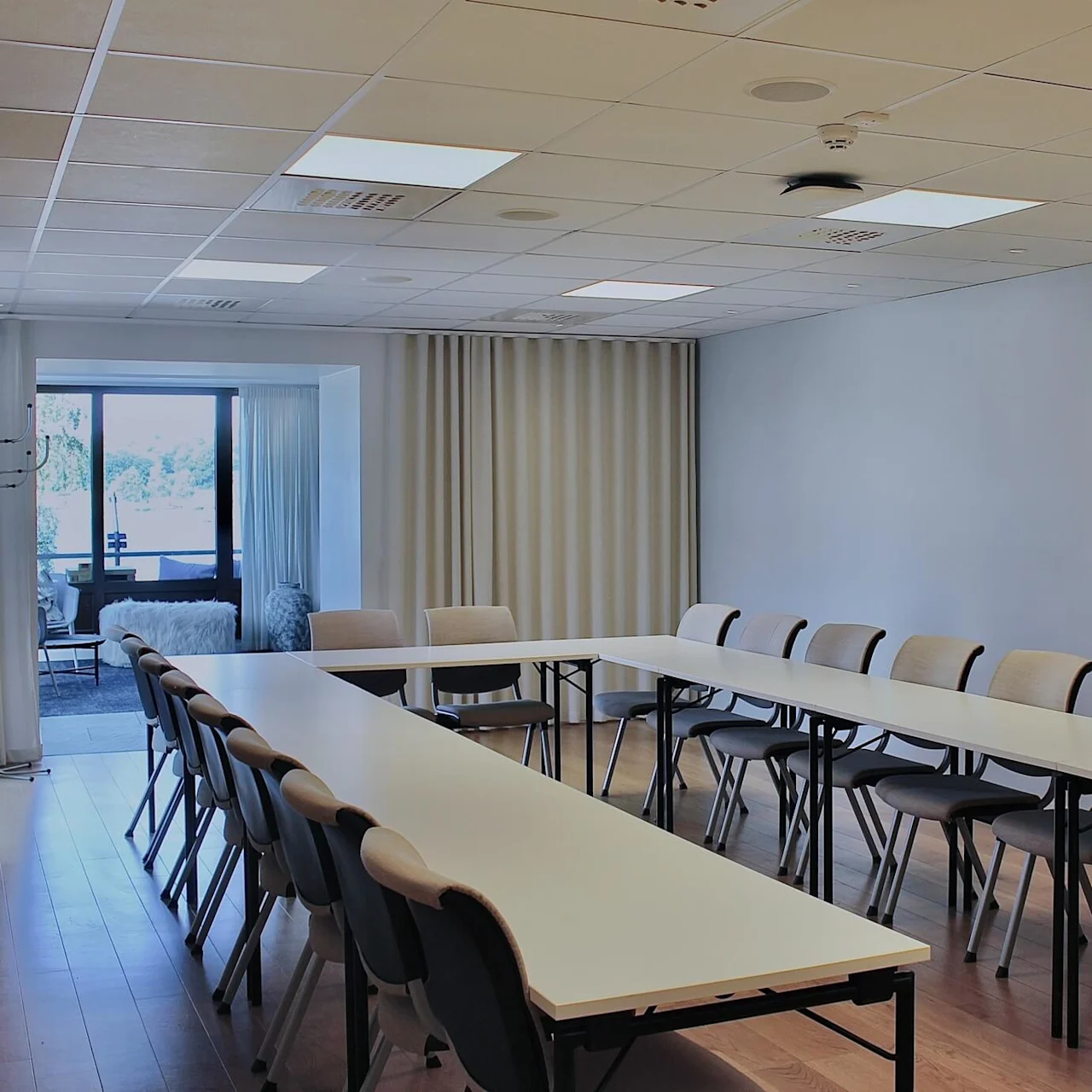 Tables form a U-shape with chairs around them in a spacious conference room, featuring wooden flooring and a large window offering a view outside.