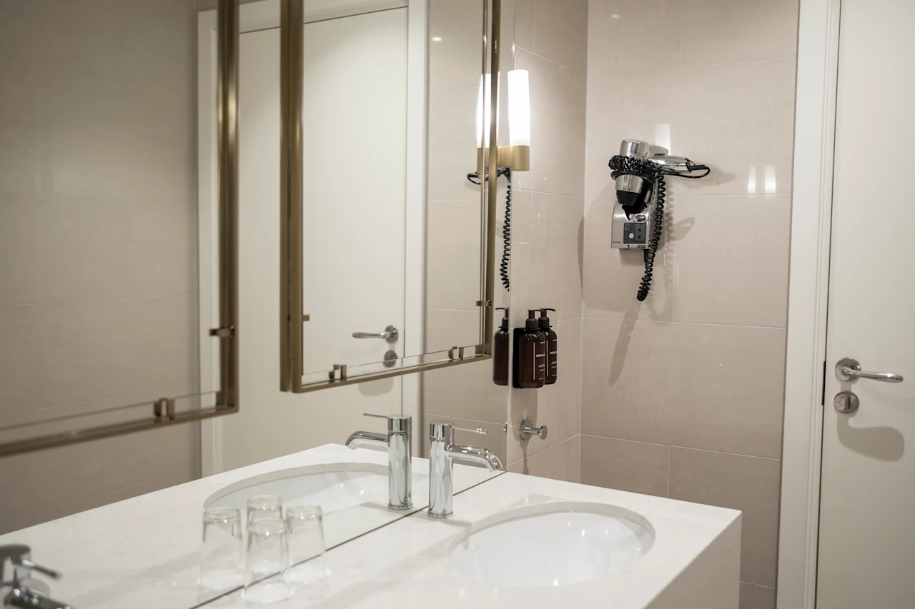 Et lyst og moderne badeværelse med dobbelt håndvask, et stort spejl, vægmonteret hårtørrer og flydende sæbedispensere.