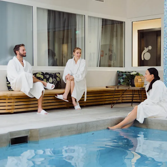 Tre personer i vita morgonrockar sitter vid en inomhuspool och pratar bredvid en kaklad pelare. De befinner sig i en spa-miljö med bänkar, kuddar och mjuk belysning.
