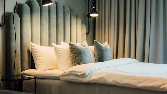 A bed with teal and white pillows rests against an upholstered headboard. Wall lamps illuminate the bed, with closed curtains in the background creating a cozy atmosphere.