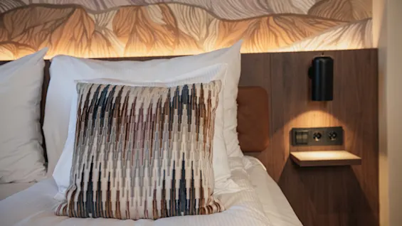 En koselig hotellseng med en dekorativ pute, myke puter og en moderne nattbordslampe med en liten hylle og stikkontakter.