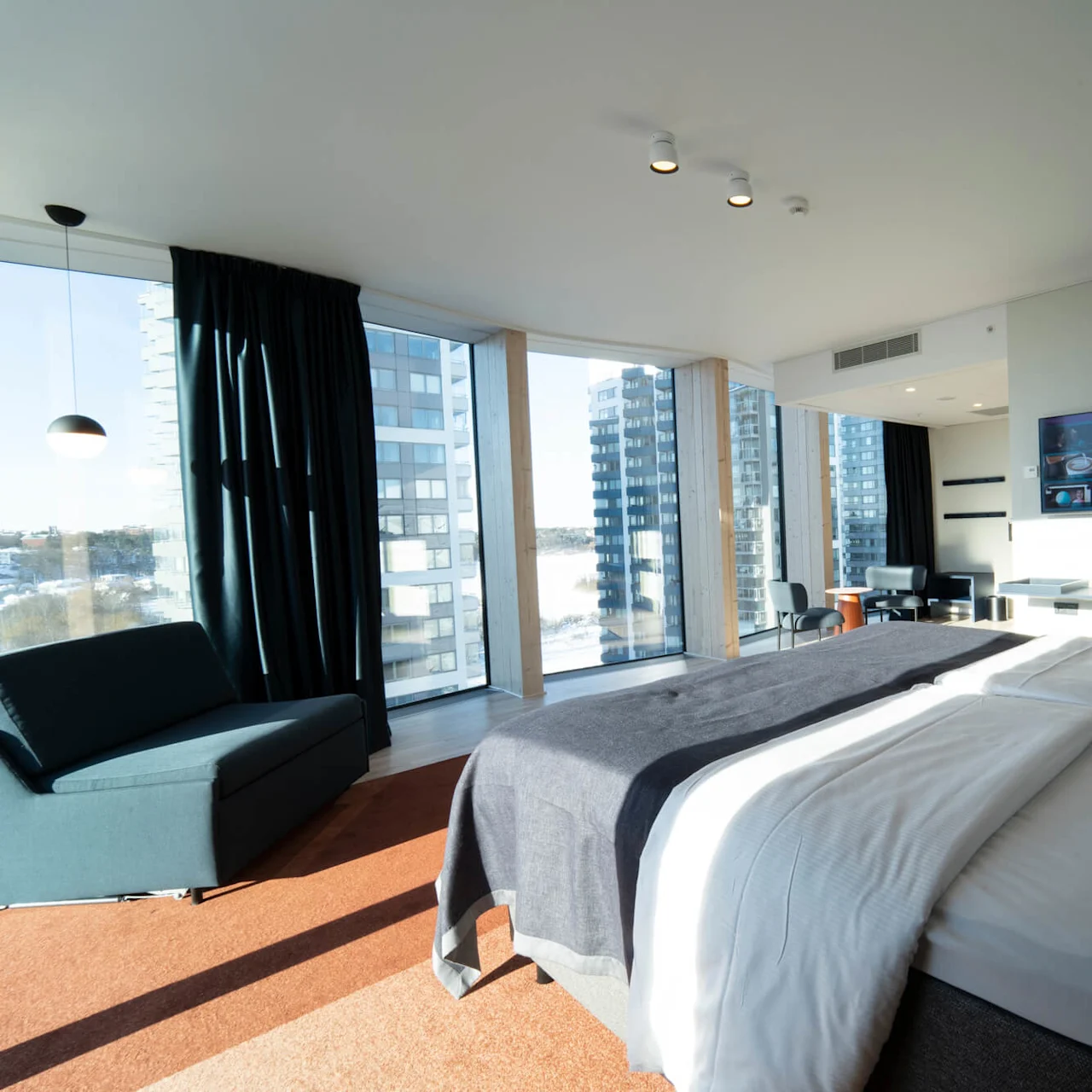 Seng med hvidt sengetøj i solrigt hotelværelse, vendt mod vægmonteret tv. Store vinduer med gardiner, sofa og stole i moderne indretning. Højhuse synlige udenfor.