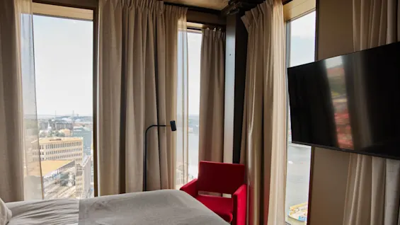 En säng med grå sängkläder står bredvid stora fönster med skira gardiner, och erbjuder utsikt över stad och vatten. En röd stol och en väggmonterad TV finns i närheten.