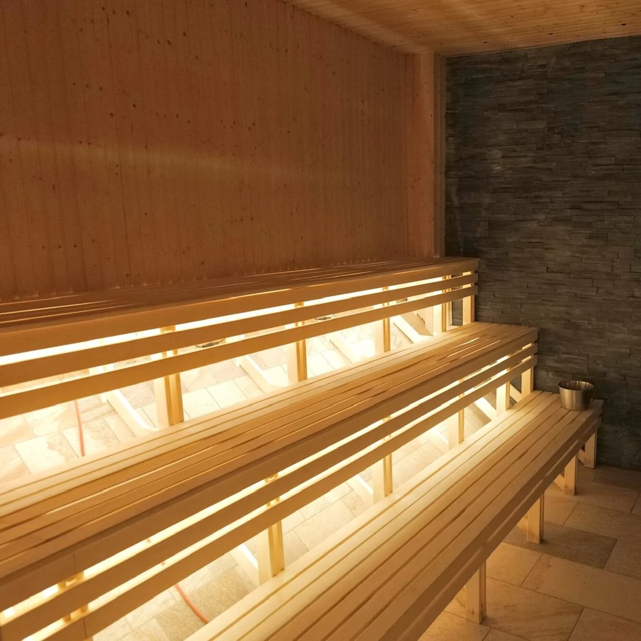 Træbænke er arrangeret i niveauer inde i en sauna, med dæmpet belysning. En stenvæg er på den ene side, og en metal kurv med sten står i hjørnet.