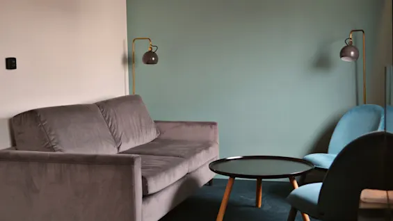 Grå sofa står stille i en minimalistisk stue med en blågrøn accentvæg, parret med et rundt sort kaffebord og to blå stole under blød belysning.