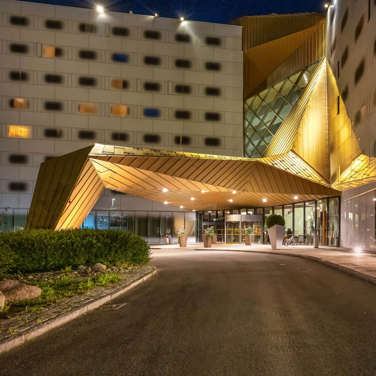 A modern building features a geometric, gold-toned entrance canopy, illuminated at night. Surrounding it are tall glass windows and symmetrical rows of square windows. Plants line the driveway.