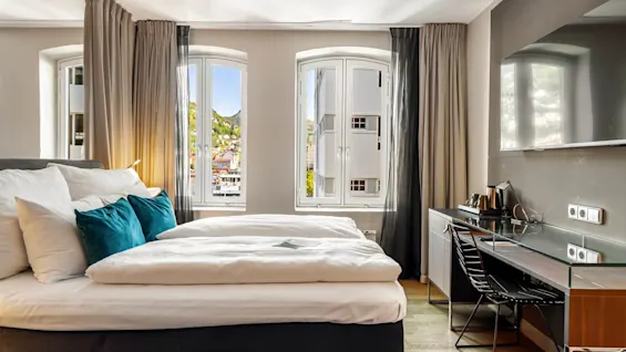 Et moderne hotelværelse har en pænt redt seng vendt mod store vinduer, med udsigt over bylandskabet. Et skrivebord med en stol, elkedel og TV fuldender den moderne indretning.