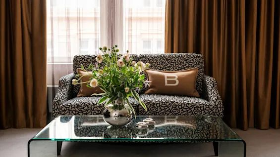 En blomstermønstret sofa har puder, én med et "B". Et glasbord foran viser en vase med blomster og to fyrfadsstager. Tunge gardiner omkranser vinduet.