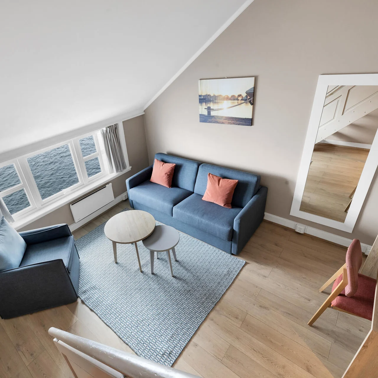 Blå sofa og stol står på et teppe mot et lite bord. Store vinduer vender mot vannet. Et speil på veggen reflekterer tregulvet. Et skrivebord med gjenstander står i nærheten.