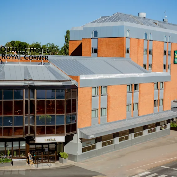 Quality Hotel med tegelröd fasad står under en klarblå himmel. Skyltar lyder "QUALITY HOTEL ROYAL CORNER" och "SOCIAL." Beläget i ett stadsmiljö med omgivande grönska.