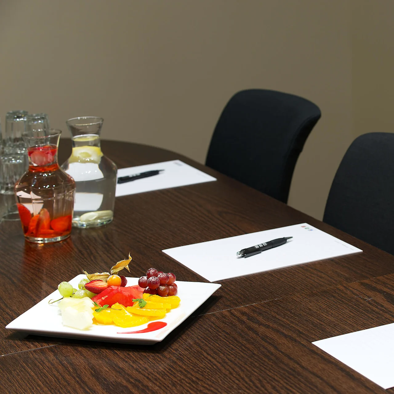A conference table set for a meeting, with papers, pens, and a fruit plate.