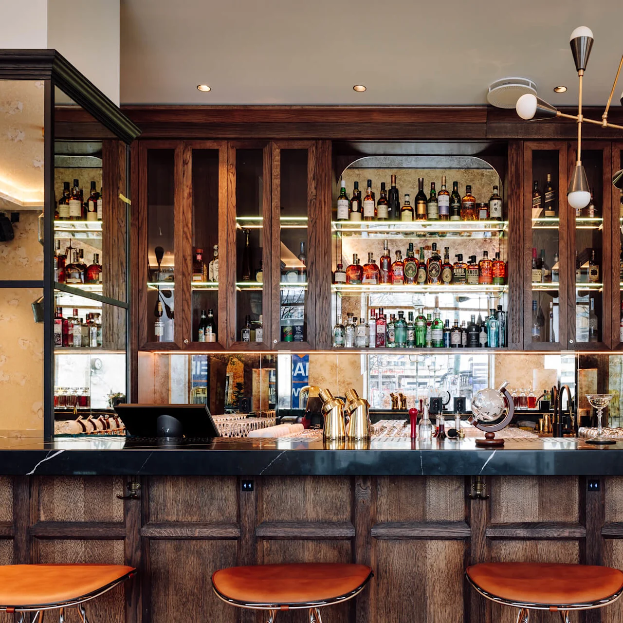 A well-stocked bar with various bottles on shelves, polished counter, and leather stools in an elegant setting. Illuminated by modern lighting.
