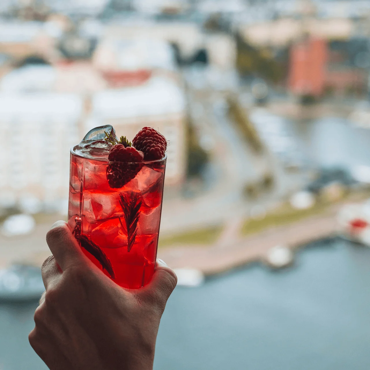 En hånd holder et klart glas med rød væske, is, hindbær og rosmarin. Baggrunden viser et uskarpt bylandskab ved vandet.