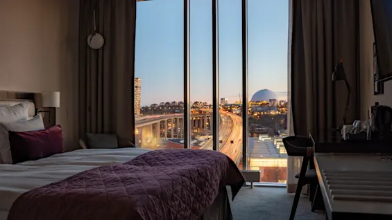 Et hotellrom med en seng med burgunderrødt sengeteppe, mot et stort vindu. Utenfor er et bylandskap synlig i skumringen, inkludert travle veier og en fjern arena.