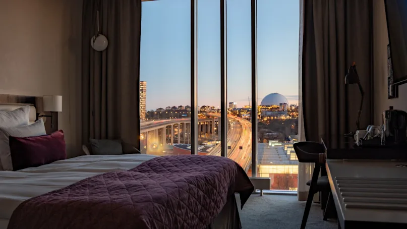 A hotel room features a bed with burgundy bedding, facing a large window. Outside, a cityscape is visible during twilight, including bustling roads and a distant arena.