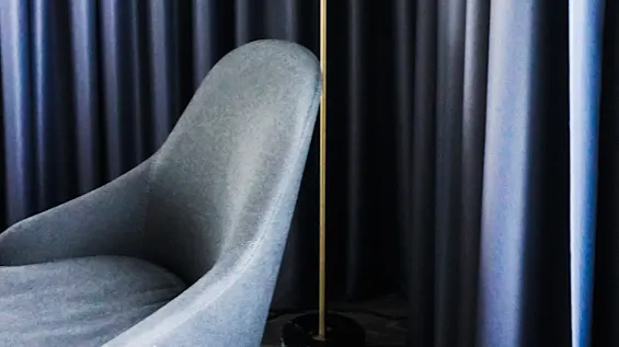 En høy gulvlampe med hvit skjerm står ved siden av en grå lenestol, mot mørke, flytende gardiner.